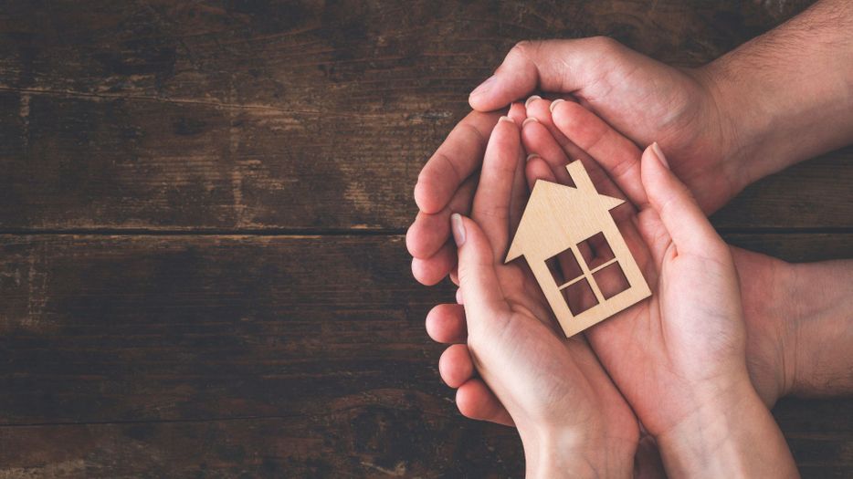 Two sets of hands laid in each other with a tiny wood house in their hands Two sets of hands laid in each other with a tiny wood house in their hands